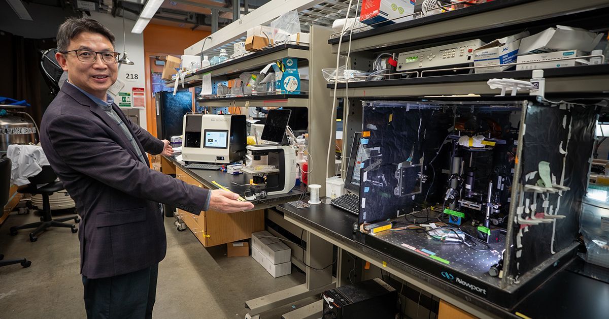 Shaochen Chen stands in a UC San Diego laboratory between an early prototype bioprinter and a later commercial bioprinter, illustrating the progression of his 3D bioprinting technology used in the ARPA-H-funded liver project.